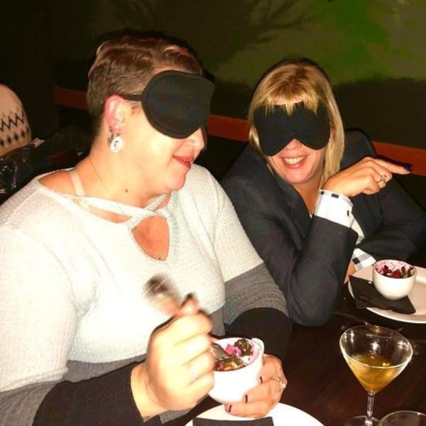 Two women wearing blindfolds, one eating from a bowl, the other smiling and pointing.