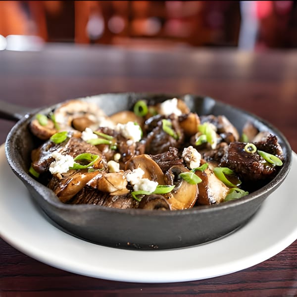 Sautéed mushrooms and steak bites topped with crumbled cheese and green onions in a cast iron skillet.