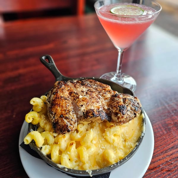 Grilled chicken over mac and cheese in a cast iron skillet, with a pink cocktail.